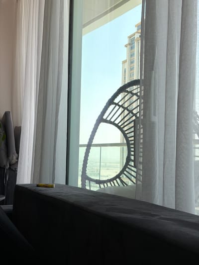 The image captures an indoor scene looking out through a window, partially obscured by curtains and furniture. In the foreground, white or light grey sheer curtains, styled like a Roman blind, frame the top and bottom of the view, creating horizontal folds. To the right, a dark grey upholstered furniture piece, likely a sofa arm or back, angles into the frame, providing a contrasting dark element. A yellow object, clearly identifiable as a "BIC" pen or lighter, is visible tucked into the side of this dark furniture.
Through the clear window, bright daylight illuminates a scene outside. A distinctive dark, woven or rattan-like circular object with radial spokes is prominent in the middle right, possibly part of an outdoor chair or decorative element on a balcony. Beyond this, a modern, multi-story building with a light-colored facade and visible windows stands under a clear, light blue sky. The scene suggests a sunny day. The location, Lusail, Qatar, implies an urban setting with contemporary architecture. No people or specific activities are depicted in the image.