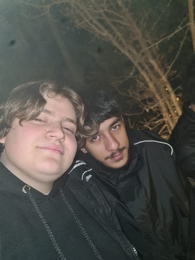 Two individuals pose closely together. One, with fair skin and wavy brown hair, is in the foreground, wearing a dark hooded sweatshirt. Behind is a person with darker skin, dark hair and a mustache, wearing a black jacket with white trim. The backdrop shows bare tree branches against a dark sky, possibly at night or in low light. The lighting suggests an outdoor setting.