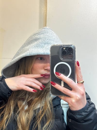 The image captures a female individual from the chest up, taking a mirror selfie in Herzliya, Israel. She is wearing a light grey hooded sweatshirt, with the hood covering most of her head, and a dark puffer jacket. Long, light brown hair extends below the hood. Her right eye is visible, looking directly towards the camera. Her lips, coated with red lipstick, are partially visible. Her left hand, adorned with red nail polish, is positioned near her mouth, with fingers touching her lower lip area. Her right hand, also with red nail polish and a gold-colored ring on the ring finger, holds a smartphone. The phone is an iPhone, encased in clear plastic, displaying a prominent MagSafe circle with an Apple logo on its back. The background consists of a plain, light-colored wall with minor vertical imperfections.