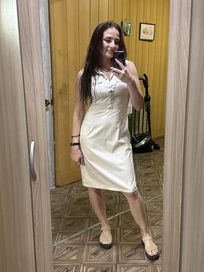 A full-length mirror reflects a young woman with long, dark hair, wearing a light-colored sleeveless dress and beige sandals. She is holding a smartphone in her right hand, taking a selfie. The background shows a room with wooden walls and a patterned tile floor. A vacuum cleaner is visible in the reflection behind her. The scene appears to be indoors, possibly in a residential setting.