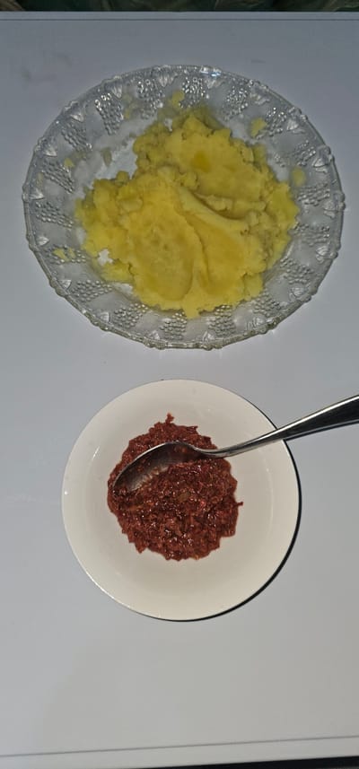 A top-down, overhead shot displays two food items on a white surface. To the left, a clear glass plate, embossed with a decorative grape vine pattern around its rim, holds a mound of fluffy, yellow mashed potatoes. Melted butter glistens on the surface of the potatoes. To the right, a white ceramic bowl contains a dark red, chunky mixture that appears to be corned beef hash. A silver spoon rests inside the bowl, its handle angled upwards. The lighting suggests an indoor setting, likely a home kitchen or dining area, possibly during the evening or in a room with artificial light. There are no visible people, text, or specific location cues beyond the general context of the Philippines, Makati. The scene depicts a simple meal presentation, with no indication of a specific event or time of day beyond the implied mealtime. The overall impression is one of everyday dining.