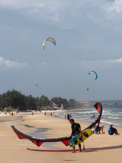 An aerial shot of a busy beach in Phan Thiet, Vietnam, shows a man in a black t-shirt walking across the sand with a yellow and blue surfboard and a large red and black kite. The kite lies across the beach in front of him. In the background, many people walk along the shoreline, and several kites are flying in the sky. Some buildings and trees are visible further inland. The ocean has choppy waves, indicating windy conditions suitable for kitesurfing.