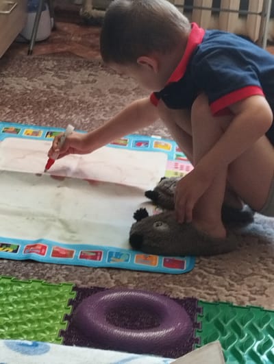 A young boy kneels on a floor, drawing on a white mat with a red marker. The boy is wearing a dark blue t-shirt with red trim and grey shorts. His feet are covered in fuzzy grey slippers shaped like animal faces. The drawing mat has a blue border with colorful images of objects. The floor is covered with a patterned carpet and puzzle mats. The background shows furniture and a door. The image was taken in Kingisepp, Russia.