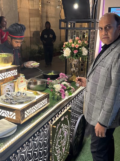 A man in a checkered coat stands near an elaborate buffet setup. The buffet features flowers, food displays, and silver serving ware. A chef in uniform is partially visible on the lower left. A figure in a black sweatshirt stands in the background. The buffet is illuminated with LED strip lights and features a decorative pattern on its surface.