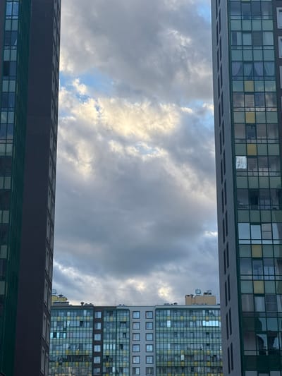 This photograph captures a view between two tall residential buildings in Murino, Russia, revealing a cloudy sky. The building on the left is a dark brown monolith with a repeating pattern of small, dark, rectangular windows. The building on the right is lighter in color, possibly gray or white, and features a grid of larger windows with green trim. Many of these windows have curtains visible, and some reflect the sky. In the distance, a lower, wider residential building is visible, also with a grid of windows, some of which are a greenish-blue color and others yellow. The sky above is filled with a dramatic cloud formation, with patches of lighter, illuminated clouds contrasting with darker, shadowed areas, suggesting it is either dawn or dusk. No people are visible in the image. The scene depicts a typical urban landscape of a modern residential district.