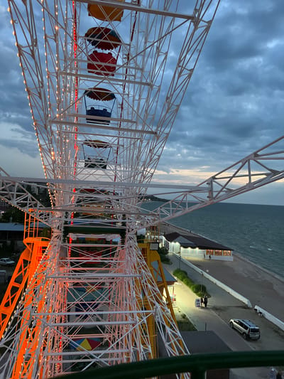 This is a vertical, eye-level shot of a Ferris wheel at dusk, likely in a seaside amusement park. The sky is filled with dark, dramatic clouds, suggesting an overcast or impending storm. The Ferris wheel structure is predominantly white, with numerous small, warm-toned lights illuminating its intricate metal framework. Some of these lights appear to be strung along the spokes and the supporting beams.
The Ferris wheel cars are visible at the top of the frame, some painted in vibrant colors like red, yellow, and blue. Below the Ferris wheel, the scene transitions to a view of a beach and the sea, the water appearing dark and choppy under the gloomy sky. Along the shoreline, there are buildings with illuminated windows, suggesting a commercial area or boardwalk. A paved path runs alongside the beach, and on this path, there are a few people and a parked silver car. The overall atmosphere is somewhat moody due to the weather and time of day, but the lights on the Ferris wheel add a touch of festive ambiance. The location is identified as the urban settlement of Shcholkine, Ukraine.