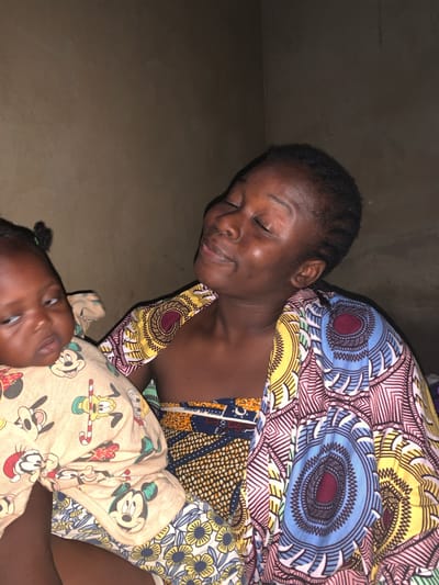 In the city of Agban, Nigeria, a young woman cradles a baby. The woman, with her eyes closed, appears to be smiling. She is wearing a vibrantly patterned cloth. The baby, whose eyes are open and looking to the side, is dressed in a light-colored garment adorned with cartoon characters, including Mickey Mouse. The background is a textured, dark brown wall.