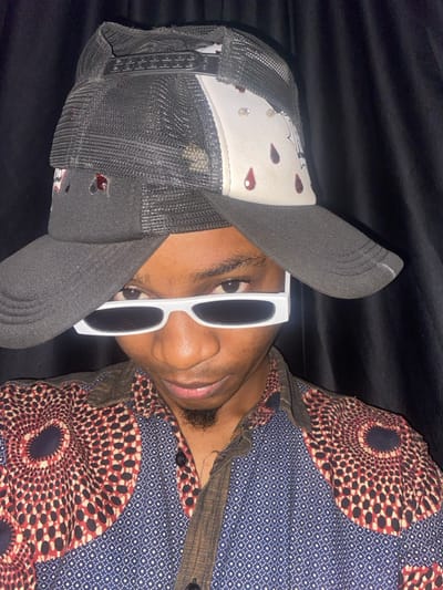 This image depicts a male individual in a close-up, chest-up shot, positioned against a dark, folded fabric background, likely a black curtain, in Ablekuma, Ghana. The subject wears two baseball caps: a dark grey or black mesh-back cap as the base, topped by a second trucker-style cap. The top cap features a black mesh back, a dark brim, and a white front panel adorned with multiple red teardrop-shaped motifs and small silver-colored studs. The individual also wears white-framed, rectangular sunglasses with dark lenses, obscuring the upper part of the eyes. A short goatee is visible on the chin. The subject is dressed in a collared, button-down shirt with a complex pattern; the main body is dark blue with a small white dot pattern, while the shoulders feature a prominent circular, radial design in red, black, and white. The collar and placket are a solid dark brown. The individual's head is slightly tilted, and the gaze is directed straight towards the camera.
