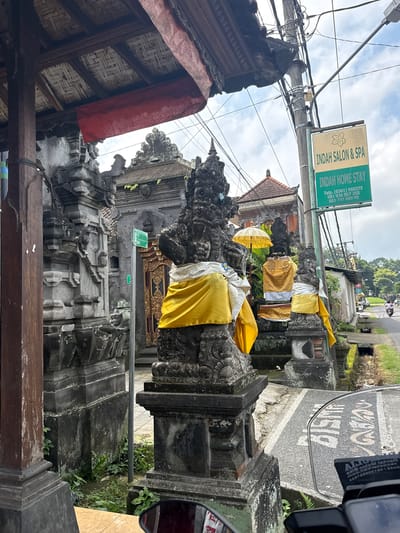 The image depicts a street scene in Payangan, Indonesia, featuring traditional Balinese statues and a sign for "INDAH SALON & SPA" and "INDAH HOME STAY." The statues, adorned with yellow and white cloths, are positioned in front of a temple-like structure with ornate stone carvings. A paved road runs alongside the property, with a small canal bordering it. In the background, trees and houses are visible, suggesting a semi-rural or suburban setting. A few motorcycles are seen on the road, indicating ongoing activity. The sky is overcast with clouds. The overall impression is one of cultural heritage integrated with local commerce.