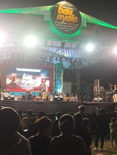 This media file captures a night scene at an outdoor festival stage, likely in Kecamatan Pujut, Indonesia. The image is taken from the perspective of the audience, looking towards the brightly lit stage.
The main subject is a large stage structure equipped with extensive lighting rigs and a prominent LED screen. Above the stage, a circular banner is suspended, displaying the text "festival pesona bau nyale 2025" with a stylized wave graphic beneath. This text is also clearly visible on the large LED screen, which shows a colorful background featuring what appears to be traditional figures or mythical characters in an artistic style, alongside the festival name and year.
On the stage, a person dressed in traditional attire, including a patterned sarong and a head covering, is standing and speaking into a microphone. This individual appears to be a performer or presenter. Other equipment, such as speakers and stage monitors, are visible on the stage floor. To the right of the stage, additional sound equipment and camera personnel with tripods suggest a professional event setup.
In the foreground, the backs of numerous audience members' heads are visible, indicating a crowd gathered to watch the event. The atmosphere is illuminated by the intense stage lights, creating bright flares and highlighting the overall dark surroundings of the night. The event is clearly taking place after sunset, as artificial lights are the primary source of illumination. The "Bau Nyale" festival name suggests a connection to the traditional Bau Nyale worm-catching festival, common in Lombok, Indonesia, which aligns with the provided location context.