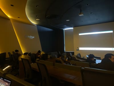 The image displays an interior view of a modern lecture hall or classroom within Universidad Anáhuac Querétaro, located in Fraccionamiento Los Héroes Querétaro, Mexico.
The environment is characterized by contrasting lighting and contemporary design. The left side features an off-white curved wall illuminated by warm yellow cove lighting, showcasing the "Anáhuac Querétaro" logo in gray. The ceiling above this section is also yellow, containing recessed spotlights. The main ceiling area is dark grey/black, equipped with multiple white recessed light fixtures, a circular air vent, and a white ceiling-mounted projector. This projector is aimed towards a large, brightly illuminated white projection screen dominating the right wall. A thin horizontal light strip runs across the wall directly beneath the screen.
The room is furnished with multiple rows of dark, ergonomic chairs and integrated dark wooden desks, arranged in a tiered, semi-circular configuration. Approximately seven individuals are visible, seated in various rows. Most individuals are positioned with their backs or sides towards the viewer, appearing to observe the illuminated screen or engage with personal items. Specific objects on desks include a yellow water bottle and several backpacks. A laptop screen is partially visible in the extreme foreground, indicating the perspective of the photographer. Individuals are seated and relatively still; no direct interactions are discernable.