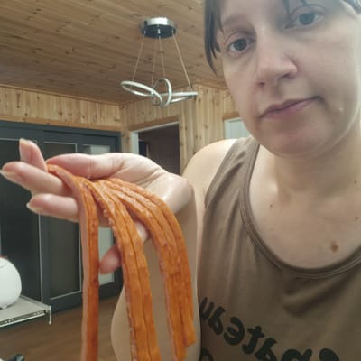 This is a selfie, likely taken indoors given the wooden wall paneling and ceiling light fixture visible in the background. The image is rotated so that the subject's face, which is partially visible at the top of the frame, appears upside down. The subject is holding a fan of sliced meat, possibly hot dogs or a similar processed meat product, in their hand, which is positioned in the lower right quadrant of the image. The meat slices are thin and have an orange-pink hue. The subject is wearing a brown tank top with visible stitching along the neckline and shoulder straps. The skin of the subject shows a few small moles. The lighting suggests it could be daytime, as there is some natural light visible. The overall scene appears casual and domestic. There is text on the subject's tank top, appearing to be a stylized font, but it is partially obscured and difficult to read fully.