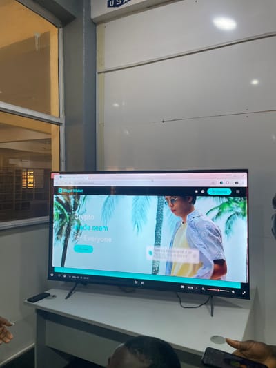 In a brightly lit room, a large flat-screen television displays the Bitget Wallet website. The website features a young man in sunglasses with palm trees in the background and the tagline "Crypto made seam for everyone." A prominent "Download" button is visible. Below the television, a white desk holds a black remote control and a smartphone being held by an individual whose hand is partially visible. The background shows a wall with some lighting fixtures. The scene suggests a technology or office environment. The reflection in the window on the left hints at an urban setting with some architectural elements. The mention of Markurdi, Nigeria, provides a specific geographical context for this observation.