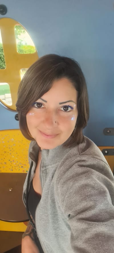 A woman is indoors, likely at a playground based on the yellow and blue structure visible in the background. She has brown hair, a nose piercing, and blue makeup with some glitter on her face. She is wearing a gray zip-up jacket over a black top. The environment seems to be brightly lit. The city of Nahariyya, Israel, cannot be confirmed from the image.