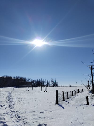 The image presents a snow-covered outdoor scene in the city of Карамыш, Russia, under a clear, bright blue sky with a prominent sun in the upper left quadrant, exhibiting significant lens flare and visible rays. The ground is entirely blanketed in sparkling snow. A clear path of human footprints extends from the lower left corner into the mid-ground. To the right, a low, chain-link fence supported by dark wooden posts runs diagonally through the snow, with small animal tracks visible adjacent to it. In the mid-ground, a flat, snow-covered field contains two metal structures resembling goalposts or playground frames, each consisting of vertical posts and a horizontal bar. A dark tire is partially buried in the snow near one structure, and a dark, upright object stands between them. A line of bare deciduous trees is visible along the distant horizon, behind which a multi-story, light-colored building is partially discernible on the far left. A utility pole with multiple cross arms and wires stands on the far right. No individuals are present in the scene.