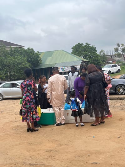 In Galadimawa Village, Nigeria, a group of people are gathered outdoors on a dirt lot near a building with a green corrugated metal roof. Several individuals, including women in dresses and a man in traditional attire, are interacting around a large green cooler and some white containers. A young girl wearing a backpack and a child in a dark dress are also present. In the background, cars are parked, including a silver sedan and a white SUV, with buildings and trees visible. The sky is overcast.