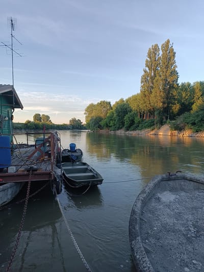 A wide, tranquil river flows through a verdant landscape under a clear, pale blue sky with scattered wispy clouds. The sun is low, casting a warm glow on the right bank, which is densely lined with trees. The left bank features a wooden dock, painted green on its structure, with various objects on it, including large blue containers and a smaller green boat with an outboard motor. Another grey, weathered boat is moored in the foreground. A tall TV antenna stands on the left side of the dock. The scene appears to be a peaceful, natural setting, likely a riverbank or marina, possibly in or near Viadana, Italy.