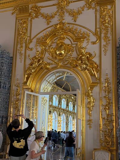In the Catherine Palace's Grand Hall in Pushkin, Russia, a wide-angle, eye-level, full shot captures the opulence of the baroque interior. The dominant feature is a magnificent, gilded archway adorned with intricate floral motifs, cherubs, and sculptural figures. Through the open doors of the archway, a long hall lined with tall windows and golden decorations is visible, reflecting bright sunlight. In the foreground, three tourists stand facing away from the camera. On the left, a young person in a black hoodie with yellow stickers on the back takes a photo with their phone. To their right, a woman in a light-colored shirt holds a tablet and appears to be taking a photo. Next to her, another person in a light-colored shirt and dark sunglasses is also holding a tablet, possibly taking a photo or using it as a guide. The walls are decorated with blue and white chinoiserie-style tiles. The scene is brightly lit, highlighting the elaborate gold decorations and the grandeur of the palace.
