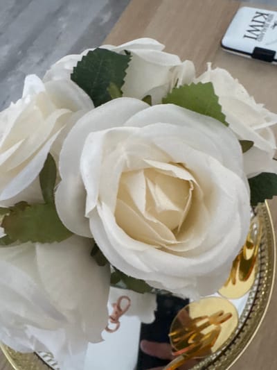 An overhead shot shows a bouquet of artificial white roses, some with visible green leaves, arranged on a circular gold decorative tray with intricate detailing along the rim. The tray also contains what appear to be gold-colored decorative elements or reflections. In the background, a light brown wooden surface is visible, and to the upper right, a white rectangular object with black lettering, possibly a wallet or cardholder, with "KIWI" and "NEW ZEALAND" printed on it. The overall scene suggests a home or display setting. There is no discernible location in Larnaca, Cyprus based on the provided image.