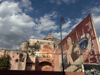 The image captures an urban street scene in Puebla City, Mexico, featuring a prominent mural on a weathered wall, old architecture, and a partially cloudy sky.
In the midground, a large, reddish-brown and pinkish-orange wall dominates the right side of the frame, angled sharply towards the viewer. This wall is adorned with a vibrant, though faded and peeling, street art mural. The mural depicts a stylized, light-skinned human-like figure with a complex, brain-like internal structure visible within its head. The figure appears to be interacting with a large, abstract, colorful object that resembles a fragmented, perhaps mechanical, device or a surreal landscape. The colors in the mural include blues, greens, yellows, and oranges, contrasting with the dark red of the wall. Above the mural, at the top right corner of the wall, the text "PENSION 24 HRS" is visible in white and black lettering. Graffiti is also present at the base of this wall, though largely illegible.
To the left of the mural, and partially obscured, stands a black lamppost with a visible light fixture. Behind the lamppost and the mural wall, a much older, decaying stone structure rises, showing signs of significant age with crumbling plaster, exposed brick, and abundant wild vegetation growing on its surfaces. At the very top of this background structure, a red-domed roof, typical of a church or colonial building, is visible against the sky. A chain-link fence runs along the bottom of the older walls.
The sky above is a mix of deep blue patches and scattered, fluffy white and grey clouds, suggesting a partly cloudy day. The overall lighting indicates daytime. The scene evokes a sense of historical depth combined with contemporary urban art, reflecting the character of an old city.