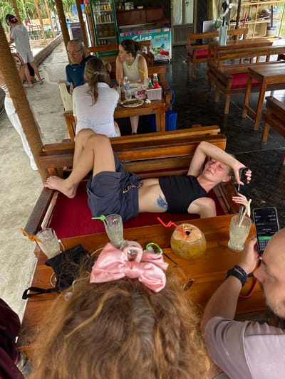 The image captures an outdoor seating area at a establishment in Nam Hoa Lư Ward, Vietnam, likely a restaurant or cafe given the presence of tables, chairs, and drinks. In the foreground, a woman is lying on a wooden bench, propped up on her arm, with a black crop top and blue shorts. Her legs are extended and bare. To her right, a coconut with a straw sits on the table. Another person, partially visible from behind, is looking at a smartphone, their arm wearing a beaded bracelet. On the table, there are two glasses with straws, a small black bag, and a pink hair bow. In the background, other patrons are seated at tables. A man with glasses is speaking to a woman wearing a white top. A refrigerator stocked with drinks is visible in the background, along with shelves holding various items. The environment appears casual and somewhat rustic, with wooden furniture and tropical foliage.