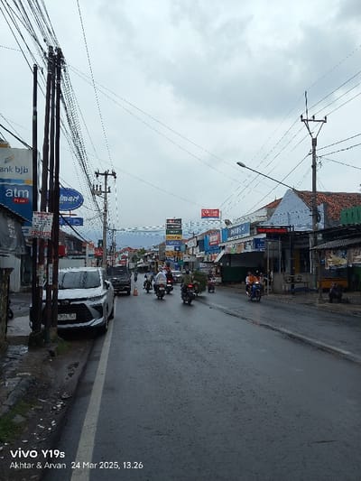 The image shows a street scene in Kecamatan Haurgeulis, Indonesia (latitude: -6.458, longitude: 107.944). The street is wet and appears to be recently rained on. The perspective is from the curb on the left side of the road. There are power lines running along the street, as well as several utility poles. On the left side of the road, there are several buildings, including a shop with a sign that says "Bank bjb atm". On the right side of the road, there are more shops and buildings, including one that says "Dan Sar" and another that says "RMPS". There are several motorbikes and a white car driving on the road. There are also several people walking and driving motorbikes in the street. The sky is overcast with gray clouds.