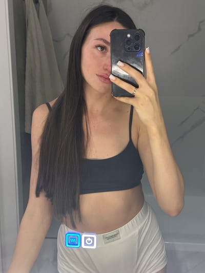 A woman with long dark hair, wearing a black sports bra and white shorts, takes a selfie in a bathroom. She is holding a black smartphone in her right hand, with her left hand resting on her hip. The background shows a tiled wall and a towel hanging on the left. Two illuminated icons, one depicting wavy lines and the other a power symbol, are visible on her shorts. The location is not specified as Эстачи, Russia.