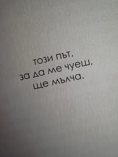 The image displays a close-up of text written on a light-colored, textured surface, likely paper. The text is in Bulgarian and is oriented diagonally from the bottom left to the top right of the frame. The words are in black, sans-serif font. The text reads: "този път, за да ме чуеш, ще мълча." which translates to "this time, so you can hear me, I will be silent." The background is a plain, uniform gray with subtle vertical textural lines, suggesting it is a document or a similar printed material. There are no people, animals, or any other objects visible in the image. The lighting appears to be ambient, creating no harsh shadows. There is no specific geographical location in Pazardzhik, Bulgaria, that can be identified from this image.