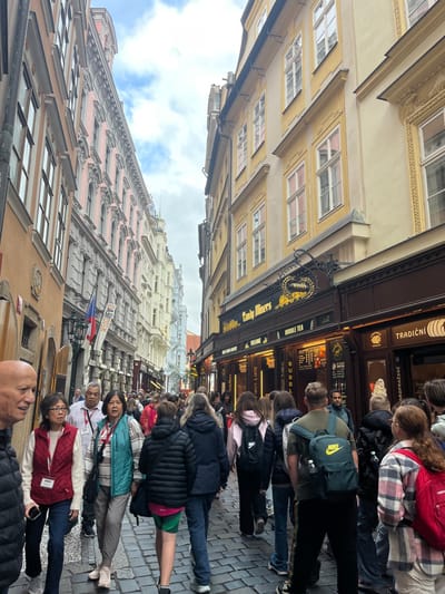 The image depicts a densely populated, narrow cobblestone street in the Capital City of Prague, Czechia, under an overcast sky. Multi-story buildings with ornate historical architecture line both sides of the thoroughfare. The left side features structures with light orange-brown and pink facades, displaying numerous windows, with a Czech flag affixed to one building. The right side is dominated by a yellow building, with its ground floor housing commercial establishments, most notably "Candy Miners," identifiable by its dark storefronts, gold lettering, and advertisements for "Bubble Tea," "Sweets & Coffee," and "Tradiční." An elaborate black wrought-iron sign with a golden emblem is mounted above the "Candy Miners" entrance.

The street is crowded with a diverse group of pedestrians, comprising individuals of various ages and ethnicities, walking in both directions. In the immediate foreground on the left, a bald Caucasian man is partially visible. Adjacent to him is an Asian woman wearing a red vest and glasses, followed by an Asian man in a white shirt with a lanyard, and another Asian woman in a teal vest. The majority of individuals further into the crowd are wearing casual clothing, with notable items including a black puffer jacket, a green Nike backpack, and a red backpack with a plaid shirt. The primary action is the general flow of pedestrian traffic navigating the crowded street.
