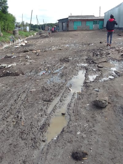 The image shows a muddy dirt road in Athi River, Kenya. The road is located at coordinates -1.465, 36.983. The road is filled with tire tracks and puddles of water. There is a large rock in the lower right corner of the image. A single person is walking away from the camera on the far right side of the image. There are several small buildings on the left side of the image. The image is taken from a low angle. The scene is deserted.