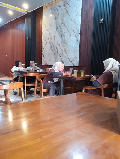 In Pandean, Indonesia, a group of four women is seated at two wooden tables in a brightly lit indoor space, possibly a cafe or restaurant. The tables are arranged such that one is in the foreground and the other slightly behind.
The woman closest to the camera, wearing a hijab and a maroon top, is using her mobile phone. To her left at the same table, another woman, also wearing a hijab, is looking at her phone. Two beverages in clear plastic cups with straws are on the table between them.
At the second table, two women are seated. One is wearing glasses and a striped shirt and appears to be writing or reading something. The other woman is wearing a hijab and a light blue top.
The walls of the space are decorated with a combination of wood paneling, vertical black stripes, and a marble-like panel with gold accents. The chairs are made of wood, matching the tables. Artificial lighting is provided by ceiling-mounted fixtures.