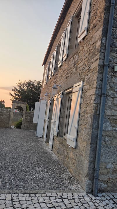 The image depicts the exterior of a stone building in Macé, France, at what appears to be dusk. The building has multiple windows with white shutters, some of which are open. A grey downspout runs vertically along the side of the building. The ground in front of the building is covered in gravel, with a cobblestone path bordering it. In the background, an arched stone structure is visible, along with trees and a hint of a sunset in the sky. There are no people visible in the image.