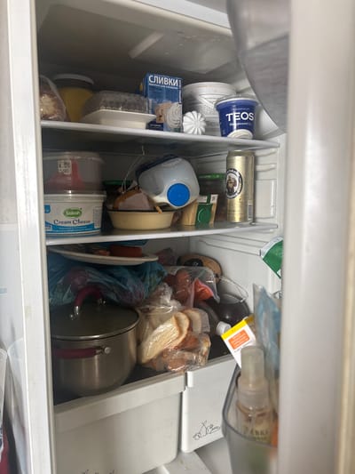 The image shows the interior of a refrigerator filled with various food items and containers. On the top shelf, there are several items including a jar of yellow liquid, a round object in a white holder, and a blue and white box labeled "СЛИВКИ WHIPPING CREAM." To the right, a white tub and a blue container labeled "TEOS ГРЕЧЕСКИЙ ЙОГУРТ 2%" are visible.

The middle shelf contains a plastic container with a red lid, a tub of "Sabah Cream Cheese," a can of beer, and a plastic bottle with a blue cap. There are also some food items in bags and a yellow bowl.

The bottom shelf is cluttered with plastic bags containing food items, including what appears to be bread and sausages. A stainless steel pot with red handles is prominently placed in the foreground. A clear plastic container with a spray bottle inside is visible on the right side of the bottom shelf. The overall impression is that of a well-used refrigerator with a variety of contents. No specific location in the city of Niva, Russia, can be determined from the image itself.