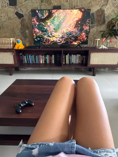 A person's legs are visible in the foreground, wearing denim shorts, as they sit facing a television screen. The television displays a colorful, abstract image. Behind the television is a stone wall. A wooden media console with shelves filled with books and decorative items is situated below the television. A basket and a yellow plush toy are placed on top of the console. A dark wooden coffee table is in front of the person, with a video game controller resting on it. The scene appears to be an indoor living room.