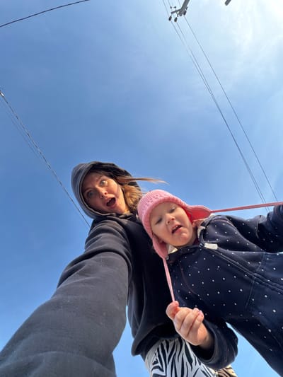 The image presents an unusual, inverted perspective of an adult and a young child looking directly down at the camera, set against a clear blue sky. The scene takes place outdoors in Gornyi (Dvurechenskoe S/P), Russia, during bright daylight hours.

The adult, positioned on the lower right, has light brown hair, which is partially covered by a dark hooded sweatshirt. Her eyes are wide, and her mouth is open, suggesting a playful or surprised expression. Her arm, clad in the dark sweatshirt, extends into the frame.

Above the adult, a young child is visible, seemingly held upside down or leaning over. The child wears a pink knitted hat with ear flaps and a dark blue jacket featuring white polka dots. A garment with a black and white striped or zebra pattern is visible beneath the jacket. The child has a wide, open-mouthed smile, revealing teeth, and appears joyful. A pink string or strap is visible near the child's chin.

The background consists primarily of an expansive, bright blue sky, indicating clear weather. Several dark utility wires traverse the sky, and a portion of a utility pole is visible on the left, hinting at an outdoor setting that could be residential or rural. The overall mood is one of lightheartedness and playfulness. There is no discernible text visible within the image.