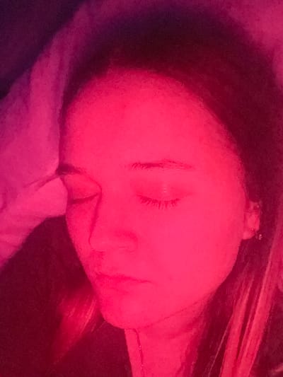 A single individual, appearing to be a female adult or young adult, is depicted in a close-up, angled view. Her eyes are closed, consistent with a state of rest or sleep. Her head rests on an unseen surface, with light-colored fabric or bedding partially covering the upper left portion of the image. Dark hair is visible on the right side of her face. The entire scene is saturated by intense deep red and magenta light, indicating a strong artificial light source. The background is indistinct and dark, characteristic of an indoor environment with minimal ambient light beyond the primary illumination. No discernible features within the image allow for the identification of a specific location in Pushchino, Russia.