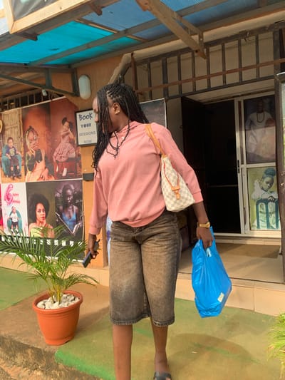 A woman is standing in front of a building with a blue plastic bag in her right hand. She is wearing a pink long-sleeved shirt and denim shorts. A white checkered purse is slung across her body. Her hair is braided and hangs down her back. She is also wearing a gold watch on her left wrist.

Behind the woman, there is a building with a glass door and windows. Inside the windows, there are framed pictures of people. There is also a sign on the wall that says "Book Your Sessions Now". Several other framed pictures are displayed on the wall, including what appears to be a photography studio's portfolio, featuring children and adults. A potted plant with green leaves is to the right of the woman.

The scene appears to be outdoors, with a greenish-brown ground surface and a blue corrugated roof structure visible on the left. The lighting suggests it could be daytime, but the overall lighting is not particularly bright, indicating it might be overcast or late in the day. The woman's posture and the way she holds the bag suggest she might be leaving or arriving at the location. The setting looks like a commercial establishment, possibly a photography studio or a shop. The overall impression is one of everyday activity.