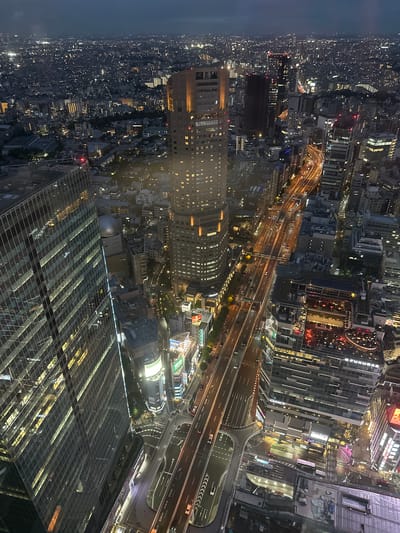 This is an aerial nighttime view of Shibuya, Japan, focusing on illuminated cityscapes. The camera is positioned high, likely within a skyscraper, offering a vantage point that reveals intricate urban details. In the foreground, the reflective glass roof of the building the camera is located in is visible.
The scene is dominated by dense urban development. Numerous high-rise buildings stand densely packed together. Interlaced highways crisscross the area. Numerous orange-colored lights indicate car traffic.
Illumination is plentiful, with buildings lit up by interior lights. The overall impression is a sprawling, vibrant urban environment after dark.