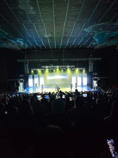 A dimly lit theater is filled with an audience watching a performance on stage. The stage is brightly lit with yellow and blue lights, illuminating several performers in black clothing. Above the stage, lighting rigs and speakers are suspended from the ceiling. The ceiling itself is dark, with a grid pattern, and appears to have a projection of clouds and stars on it, suggesting a creative stage design.

The audience is seated in rows, with most of their faces obscured by shadows. Many people are holding up their phones, likely to record or take pictures of the event. The overall atmosphere appears to be one of engagement and entertainment. The lighting suggests it is nighttime, and the indoor setting removes any external weather factors. The location context of Mykolaiv, Ukraine, provides a specific geographical setting for this event. There is no visible text in the image.