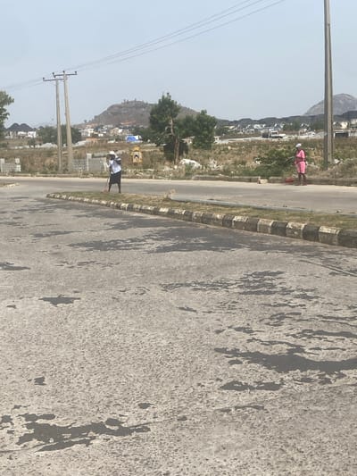 The image shows a partially cracked asphalt road in Iddo Tudunwada, Nigeria. Two people are visible, one in pink clothes and one in a white shirt and black shorts. The surrounding environment includes dry, grassy land, scattered buildings, and utility poles. A hill is visible in the background.