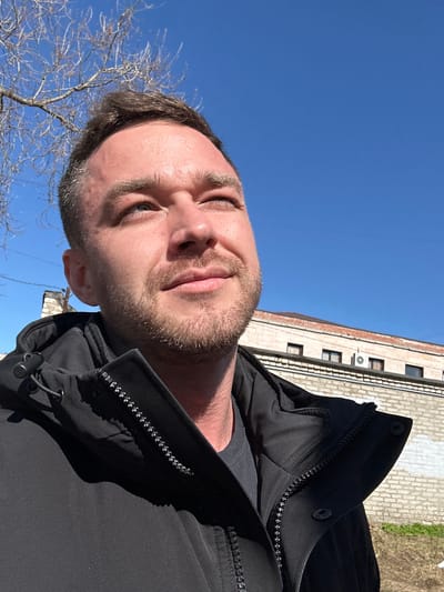 A man is pictured in a low-angle selfie outdoors. He is wearing a black jacket. Behind him is a blue sky and a building, which might place the location in an urban setting. There are bare branches of a tree visible. The exact location within Dobryanka, Russia is not discernible from this image.