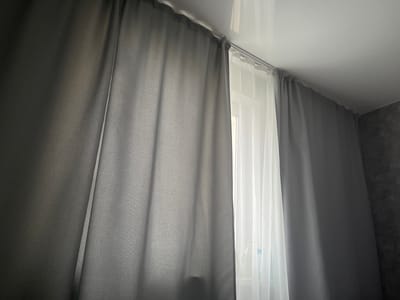 A pair of gray curtains are drawn across a window, with a sheer white curtain visible behind them. The gray curtains are thick and opaque, obscuring most of the view outside. The white curtain is thin and translucent, allowing some light to filter through. The curtains are hung from a metal rod attached to the ceiling. The room appears to be dimly lit, with natural light coming from the window. The wall to the right of the curtains is textured and dark gray. The overall impression is one of a domestic interior, likely a bedroom or living room. The scene is static and there are no people or animals visible. There is no identifiable information within the image to suggest the specific location is Kamskiye Polyani, Russia.