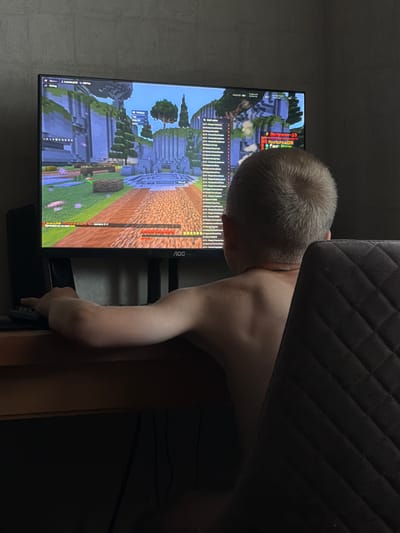 A young boy, seen from behind, sits at a desk playing a video game on a computer monitor. The boy has light brown hair and is wearing a necklace. His left arm is extended, resting on a keyboard. The monitor displays a blocky, pixelated game environment. A chair with a quilted back is visible to the right of the boy. The scene is dimly lit, suggesting it is indoors, possibly at night. There is no visible indication of the city of Vilshany, Ukraine.