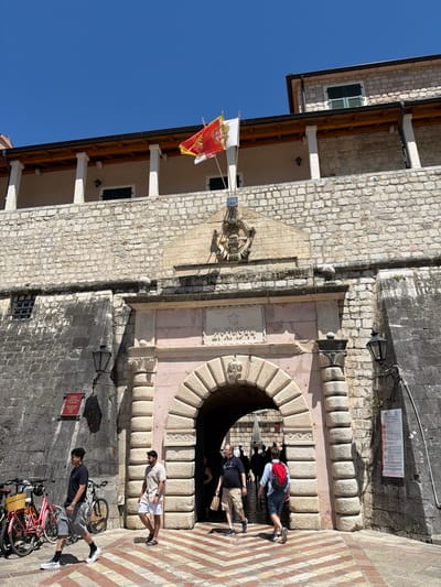 The image shows the Sea Gate in Kotor, Montenegro, on a sunny day. Two flags fly above the gate: one red and yellow with a crest, and another white flag with a crest. The gate itself is an arched passage made of weathered stone. Above the arch, an inscription in stone reads "21-XI-1944". Several people are walking through the gate. On the left side of the image, two men are walking, and a woman is visible further in the background. On the right side, two men are walking out of the gate, one wearing a red backpack. Bicycles are parked to the left of the gate, one pink with a basket. The surrounding architecture is of old stone buildings. The ground in front of the gate has a patterned walkway of red and white stones.