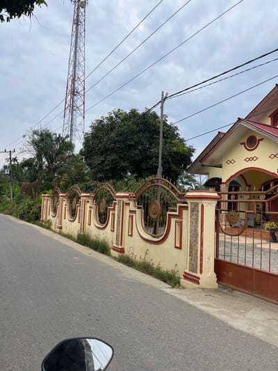 The image shows a residential street scene in Tanahterban, Indonesia, featuring a long, ornate fence along the side of the road. The fence is constructed with alternating sections of light yellow plaster and red-trimmed ornamental pillars, punctuated by decorative metalwork panels. Beyond the fence, a light yellow house with a red roof and red trim is visible. Behind the house and fence, lush green trees and foliage create a dense backdrop. In the distance, a tall, red and white lattice tower, likely a telecommunications tower, stands against a cloudy sky. Overhead, several power lines crisscross the frame. The foreground shows a portion of an asphalt road and the reflection in a motorcycle's side mirror. The scene is devoid of people.