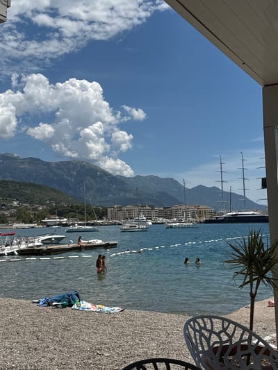 The image depicts a scenic view of a marina in Tivat, Montenegro, on a bright, sunny day with scattered clouds. The foreground shows a pebble beach with towels and beach gear laid out, suggesting a recreational area. In the mid-ground, the calm blue water of the bay is dotted with various boats, including several sailboats and a very large, dark-hulled yacht with multiple masts. A wooden pier extends into the water, where a few people are visible, some sitting on the edge and others wading in the shallow water. Further out in the bay, more vessels are anchored or moored. In the background, lush green hills rise, forming a backdrop to the marina, and beyond them, majestic mountains with rocky peaks are silhouetted against the clear sky. Part of a modern building structure is visible in the right foreground, framing the view and suggesting a waterfront establishment.