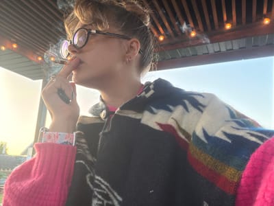 The image presents a close-up, side profile of a person, likely female, smoking a cigarette outdoors during what appears to be late afternoon or early evening, indicated by the warm, golden light. The person has light brown hair styled in a bun and wears prominent, round, dark-rimmed glasses. Visible on the ear are two distinct earrings: a silver hoop and a pink flower-shaped stud. A silver ring is on the finger holding the lit cigarette, from which smoke curls upwards. The hand also holds a small, dark object, possibly a lighter or a vape pen.
The person is dressed in a dark poncho or shawl featuring a colorful geometric pattern with white, blue, red, and yellow elements on the shoulder. Beneath this, a vibrant, textured pink long-sleeved garment with a lighter floral patterned cuff is visible.
The background features a wooden slatted roof or ceiling structure adorned with several glowing string lights. Above and to the right, a clear, bright sky is visible. On the far left, hints of distant structures, possibly a house roof, are bathed in the warm, setting sunlight. The scene suggests a relaxed, semi-outdoor setting, possibly a patio or balcony in Большая Шильна, Russia. There is no visible text in the image.