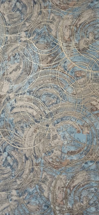 The image displays a close-up view of a textured surface, likely wallpaper or fabric, featuring an abstract pattern of overlapping circles and swirls. The colors are predominantly muted blues, grays, and earthy browns, with thin, shimmering silver lines defining the circular motifs. The texture appears somewhat rough or distressed, with variations in color and shading suggesting a layered or marbled effect. There are no discernible people, objects, or scenes. The lighting is even, with no indication of time of day or weather. The location context of Asbest, Russia, does not provide any visual cues within the image itself. No text is visible.