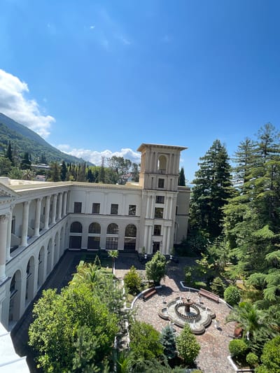 This is an exterior, high-angle, daytime shot of a grand, white, neoclassical building in Gagra, Abkhazia. The building features a long colonnade with numerous arches supported by columns. To the right of the main building, a multi-story tower with arched openings and classical detailing rises. In front of the building is a courtyard with a brick-patterned floor. In the center of the courtyard is a decorative fountain with water spraying upwards. Benches are placed around the fountain. Lush green trees, including tall evergreen trees and smaller bushes, surround the courtyard and the building. In the background, a verdant, tree-covered mountain slope with scattered houses rises under a bright blue sky with scattered white clouds. The scene is serene and well-maintained, suggesting a resort or historical landmark. There are no visible people in the image.