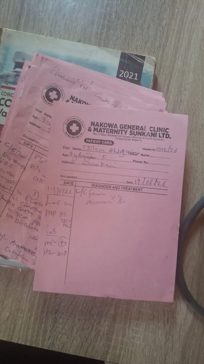 This media file shows a close-up, overhead shot of a stack of pink medical patient cards, likely from a clinic in Nigeria. The cards are arranged at an angle on a light brown, wood-grained surface. The setting appears to be indoors, with no natural light visible. The cards are the primary subject, with visible text indicating they belong to "NAKOWA GENERAL CLINIC & MATERNITY SUNKANI LTD." and are located in Sunkani, Taraba State, Nigeria.

Several cards are partially visible, but one in the foreground is the most legible. It contains handwritten entries for patient information such as "First Name," "Age," "Address," and "Occupation." There are also sections for "DATE" and "DIAGNOSIS AND TREATMENT," with handwritten notes detailing medical information. Some of the visible text includes dates like "6/6/2016" and "19/07/26," along with medical terms and prescriptions. A book with the year "2021" printed on its spine is visible peeking out from underneath the stack of cards.

The overall impression is one of official medical record-keeping. The handwriting suggests a recent or ongoing medical consultation or series of appointments. There are no people or specific actions depicted, only the documentation of medical care. The lighting is somewhat harsh, casting slight shadows, and the focus is primarily on the textured surface and the pink paper of the cards.